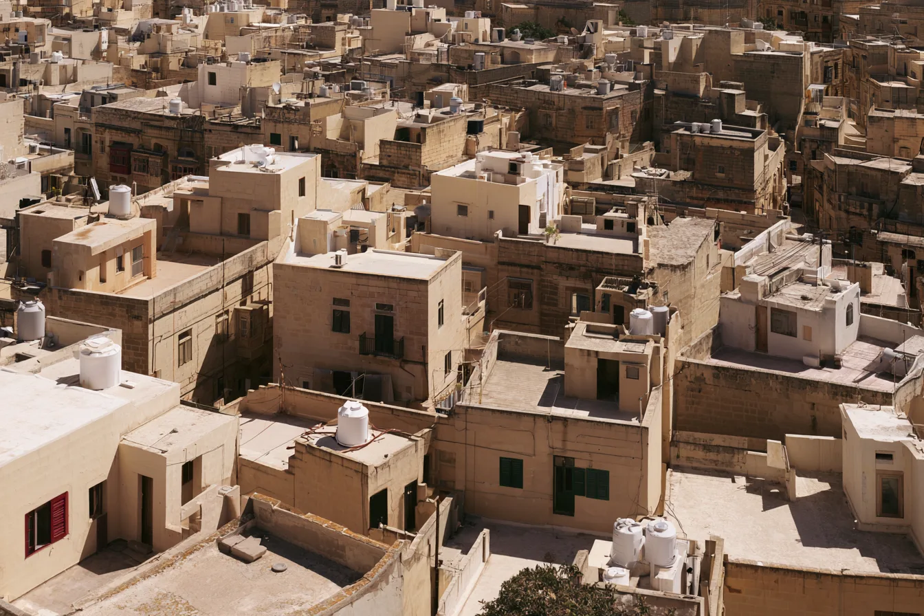 Dense Maltese townscape of flat limestone rooftops, white water tanks and green shutters under hard midday light.